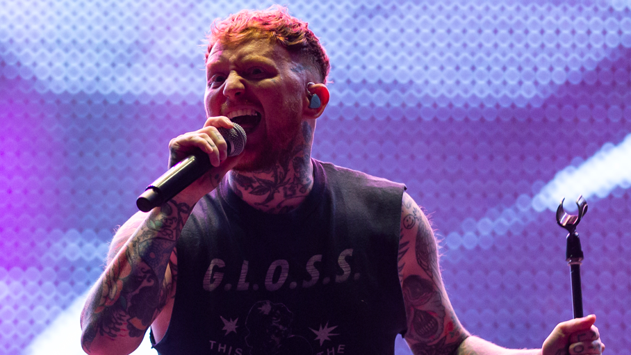 Frank Carter holding a microphone and singing to a crowd at a gig. His arms are covered in tattoos and he wears a tank-top with the word Gloss across the front.
