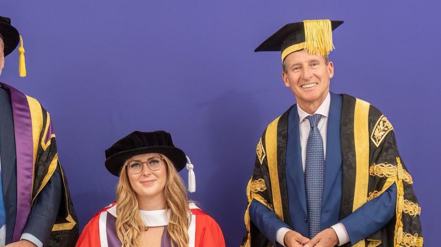 Jessica-Jane Applegate poses with Sebastian Coe in her academic dress. Applegate is sat down, wearing a red robe with a black hat and Coe is stood up wearing a black and gold robe and black mortarboard. 