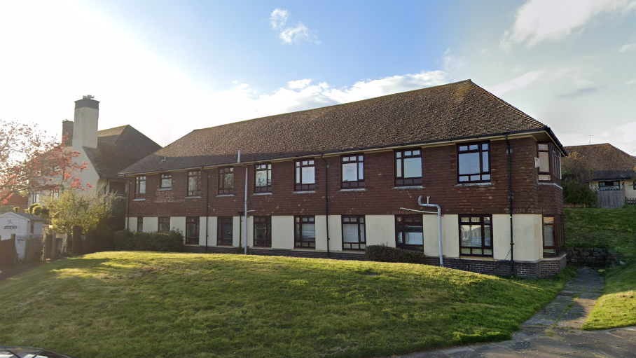 A long terraced building. The bottom half is cream the top is brown. There are 18 windows along the building. It has one storey and a dark brown roof. There is grass in front of the building and blue, cloudy sky above.