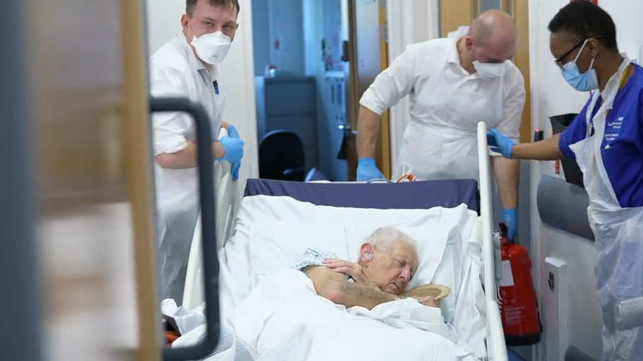 Hospital staff wheel Percy, clutching a sick bowl in his trolley, down a corridor