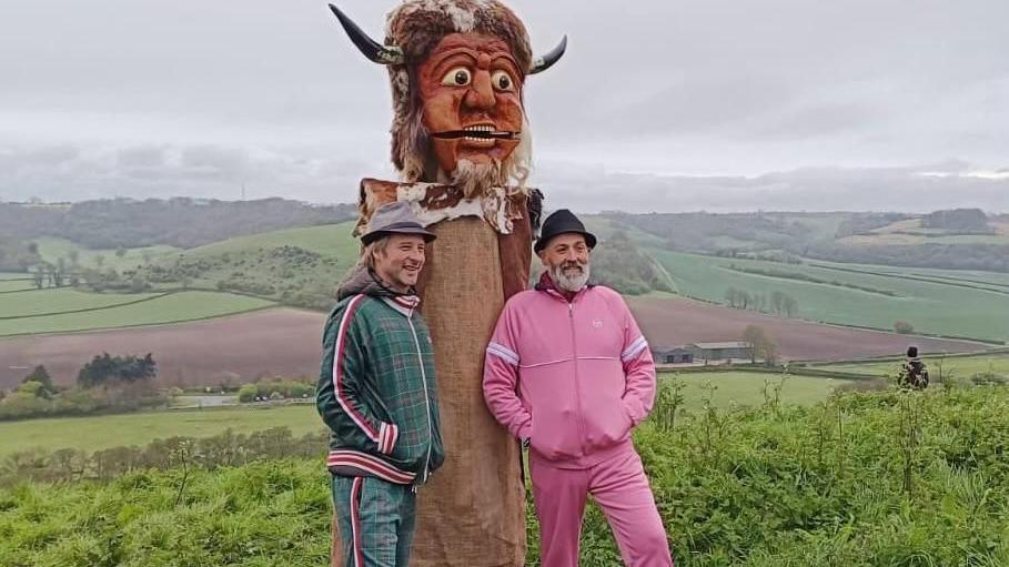 Chesney Hawkes and Vic Irvine stand either side of the Dorset Ooser. They are surrounded by countryside and up on a hill. Chesney stands to the left, wearing a green and red tartan print tracksuit, smiling as he looks at a camera to the right of the frame, and has a brown hooded puffer jacket on under his tracksuit with a grey trilby style hat. Vic Irvine stands to the right, he has a grey beard and wears a black trilby hat and bright pink tracksuit set. In the middle of the pair is the Dorset Ooser, a wooden head from 19th century folk culture. The head has a humanoid face with horns, a beard, and a hinged jaw which allows the mouth to open and close. 