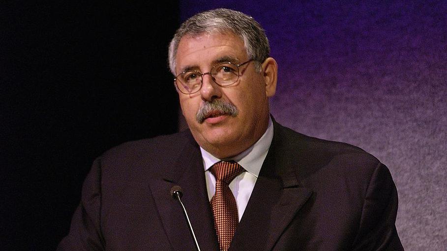 Millard Ochs, with grey hair and moustache, is stood in front of a tiny microphone dressed in a suit