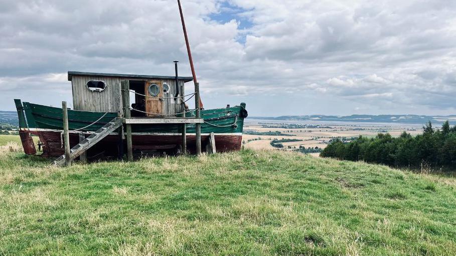 A small wooden ship structure on top of a hill. The ship has a green hull, a wooden deck, and a light coloured cabin upper section. It is sitting on a grassy field overlooking green trees a light-coloured farmland.