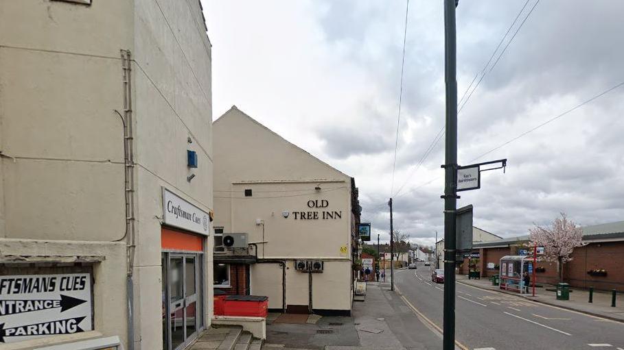 The pub in Kippax is located opposite a bus stop. The building is painted in a cream colour and lettering on the side wall reads "Old Tree Inn". A building supplies shop can be found next to the pub.