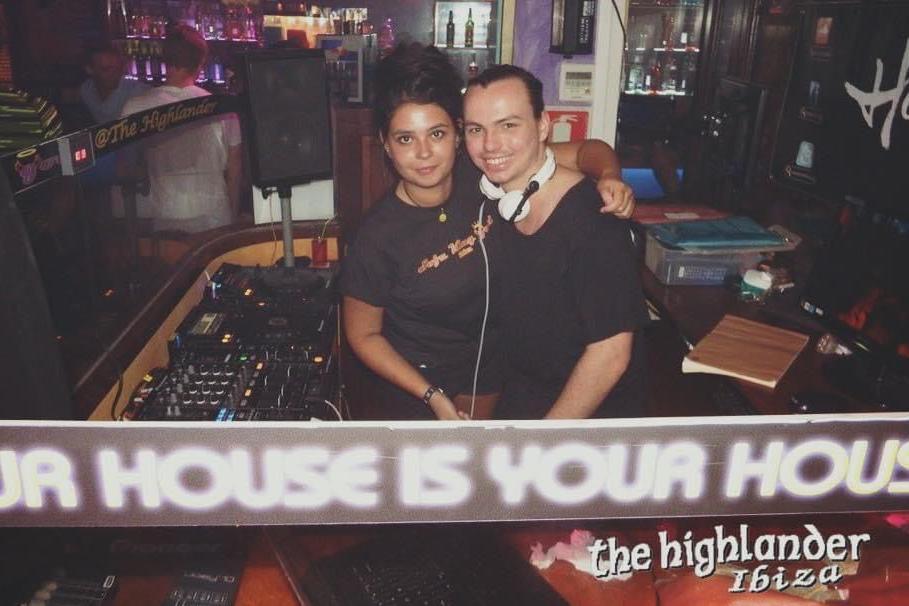 A girl in a black top with black hair has her arm around a man wearing a black top and white headphones. They both stand in a DJ booth in a bar.