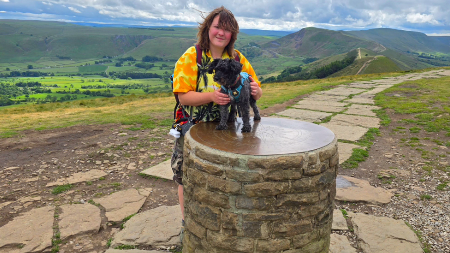 A woman wearing a bright yellow and orange shirt, with her hands on a small black dog, which is stood on a brick circular plinth. Rolling green hills can be seen in the background.