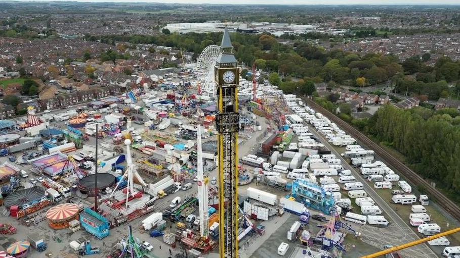 A drone image of Hull fair in construction featuring the Big Ben Tower. The tower is by far the tallest ride. It is bright yellow and is a long thin stick with a golden shiny clock tower right at the top, similar to the Big Ben clock on the Houses of Parliament.