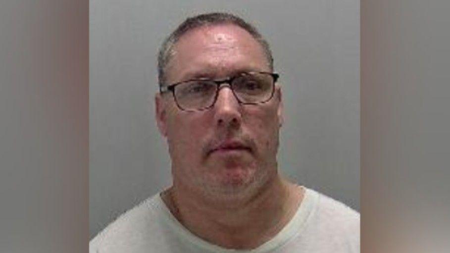 A mugshot of Scott Dobson-Smith wearing dark glasses and a white t-shirt with a grey background