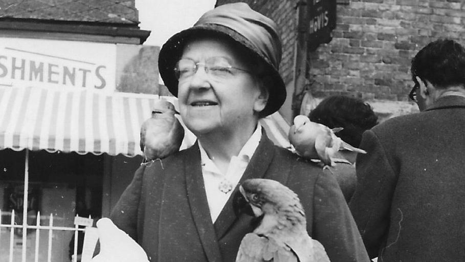 Black and white image of Catherine Brown. She is older and and wearing glasses, a hat and blazer. She has two small birds on her shoulders, and two much larger birds on her hands. She appears to be on a reasonably busy street, with shops behind her.