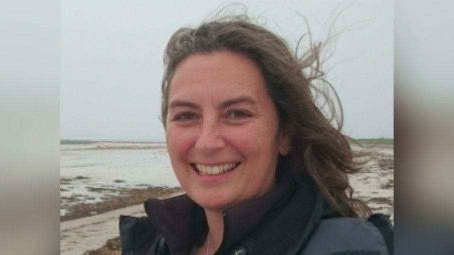 Wendy Carter with windswept long brown hair is wearing a black bodywarmer and is stood on a cloudy beach.
