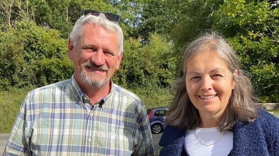 Councillors John Rudge and Sarah Murray - a grey bearded man on the left hand side is smiling and wearing a blue and green checked shirt. A woman is standing on the right hand side and is also smiling. she is wearing a white shirt underneath a textured blue coat and smiling.