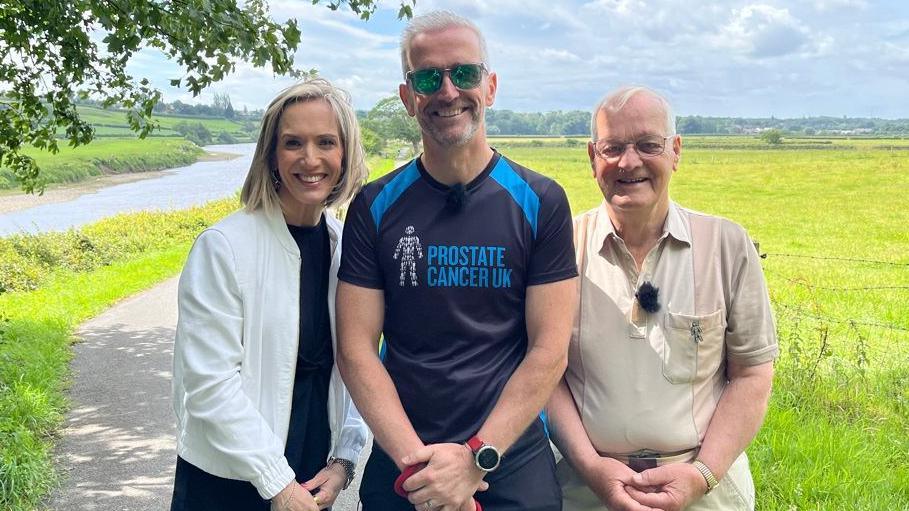 Michael Lilley is standing next to his father John and BBC North West Tonight's weather presenter Kay Crewdson on a path running through a field. He is wearing a T-shirt which says "Prostate Cancer UK".