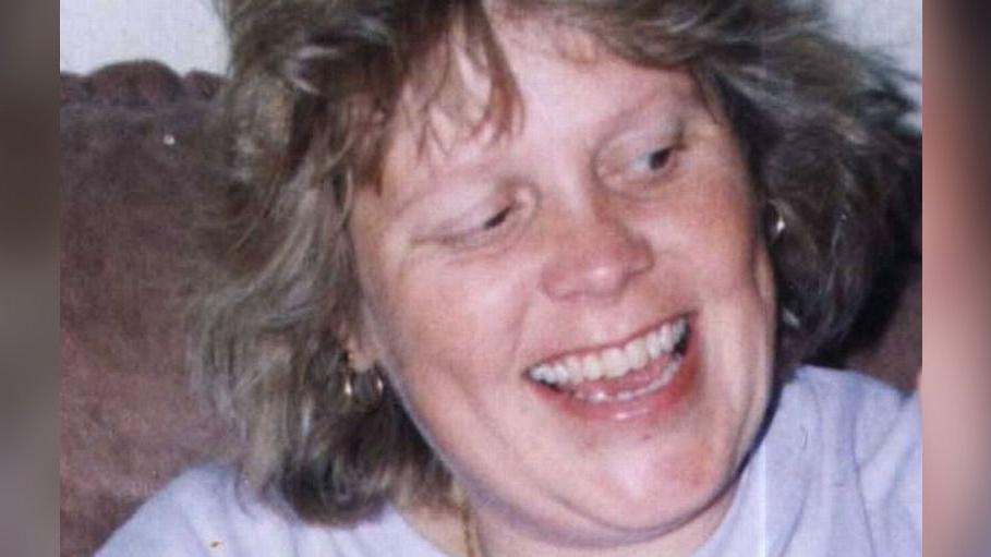 Debbie Griggs looks off camera and is smiling. She is wearing a pale coloured t-shirt and has short light brown hair.