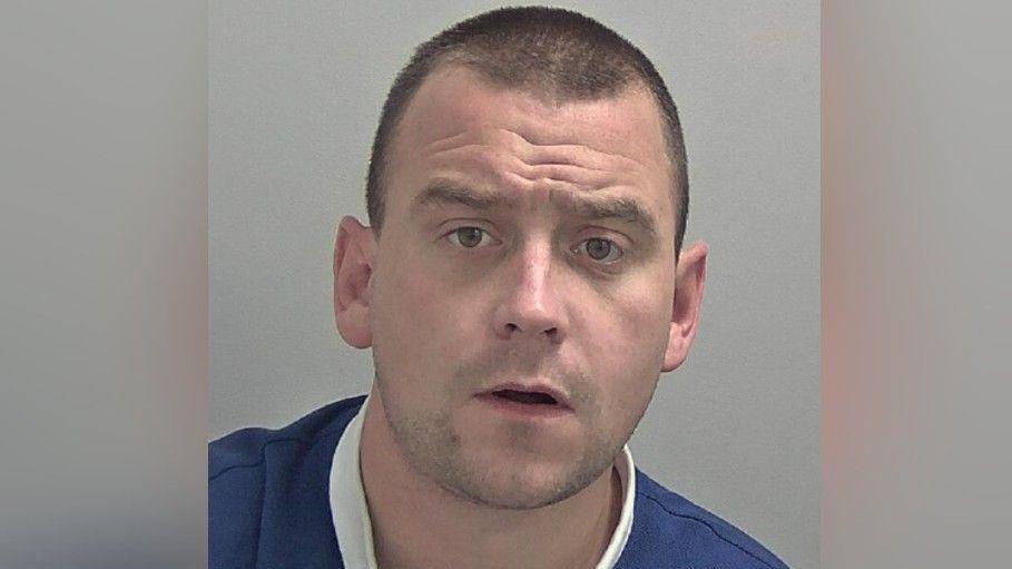 A mugshot of a man, Jakob Walpole, with short brown hair and wearing a blue and white T-shirt.
