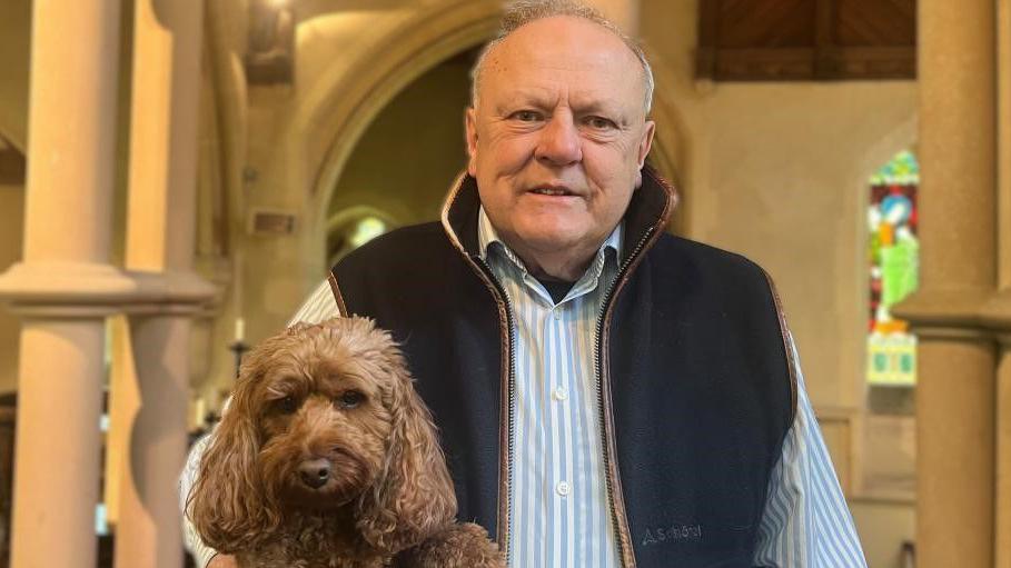 Lance Sharpus-Jones is looking into the camera, holding his dog under his arm. He is smiling and wearing a blue and white striped shirt with a sleeveless navy bodywarmer over it. His dog is a small brown cockerpoo. There are pillars on either side of him, and a stained glass window behind him to the right of the image.