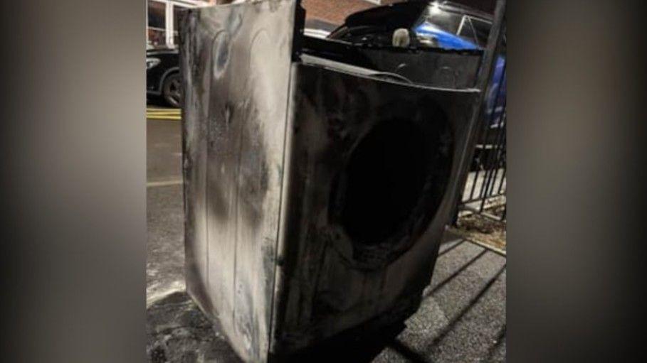 A tumble dryer which has been blackened by a fire on the pavement on the side of a road