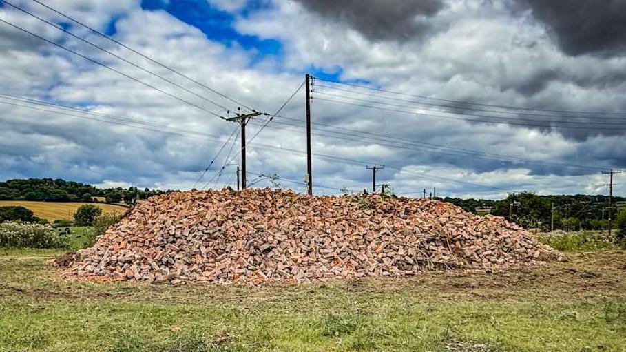 A massive pile of bricks in a field with telegraph poles behind. There are clouds in the blue sky.