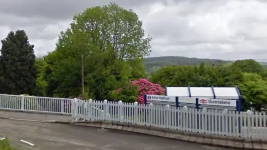 Street view of the Gunnislake train station. There is a white picket fence with a sign with three white sections. On the right it says information and on the left it says Gunnislake. Behind is trees with pink flowers