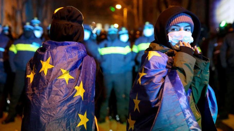 Two protesters draped in EU flags face a cordon of Georgian police wearing uniforms