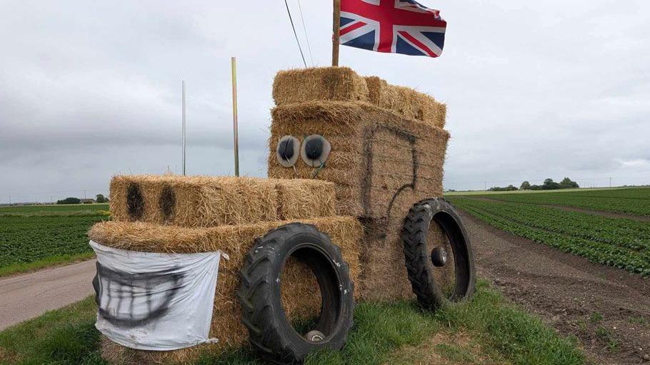 A tractor sculpture made out of straw bales. It depicts a tractor with a Union flag flying from its "cab". Two large black tyres are resting on its side. It has a face made from eyes where its windscreen would be, nostrils at the front of its bonnet and a banner painted in the shape of a smile where its bonnet grille would be. It is surrounded by fields.