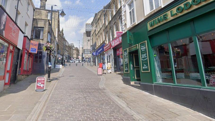 A mostly empty city centre shopping street. Shops are on both sides of the pavement, which runs uphill.