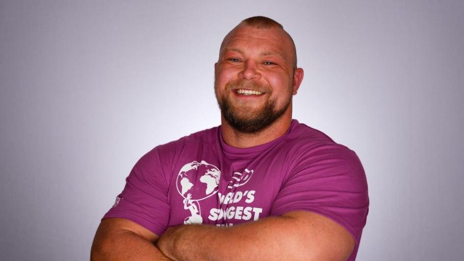 A smiling Kane Francis, wearing a purple World's Strongest Man t-shirt. He has a beard and short cropped hear and has his arms folded.