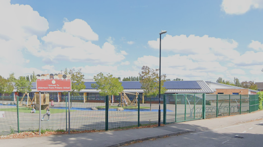 An animated image of Aldermoor Farm Primary School, which includes a playground surrounded by a green metal fence. A road and pavement run alongside.