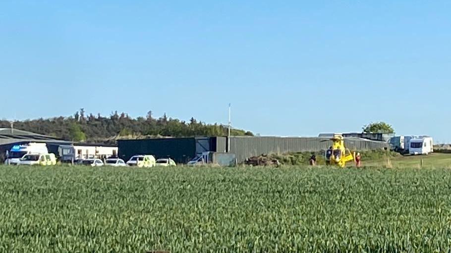Several emergency service vehicles including an ambulance helicopter in the middle of a field. Campervans can be seen in the background