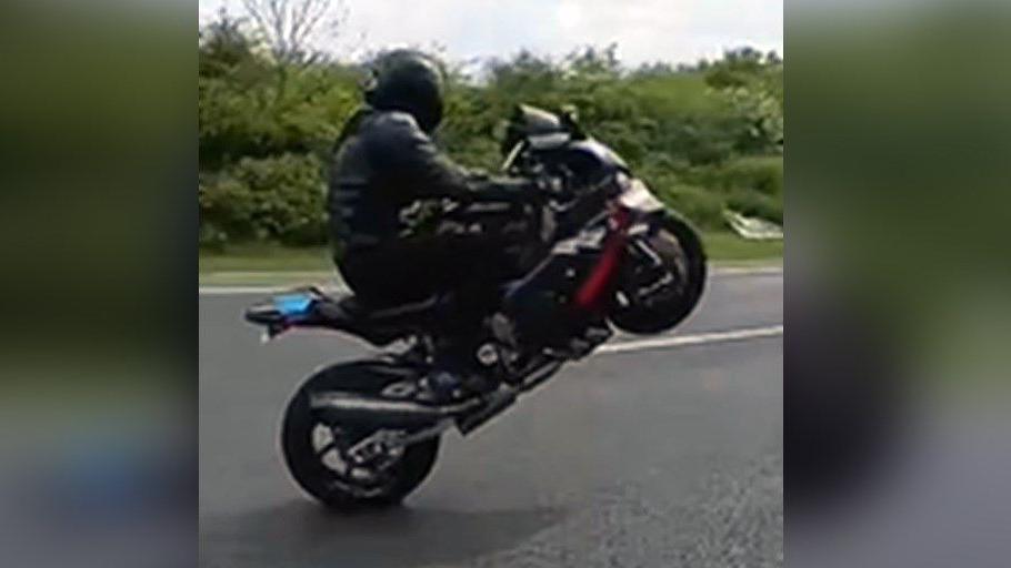 A motorcyclist on his machine wearing black leather and a black helmet. The bike they are on is also black and the front wheel is off the ground as they perform a "wheelie".