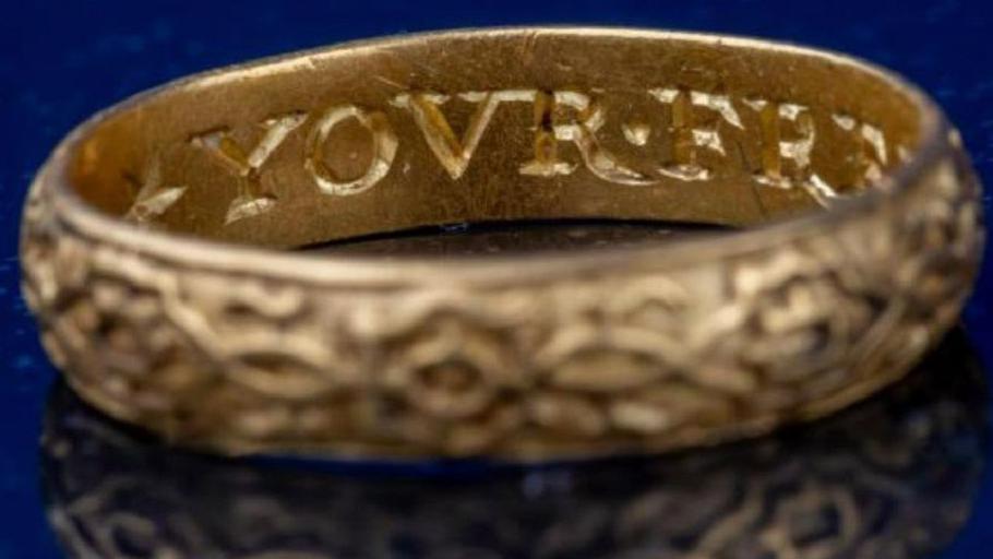A gold ring with letters carved on the inside and a pattern running along the outside. It is placed on a dark blue background. 