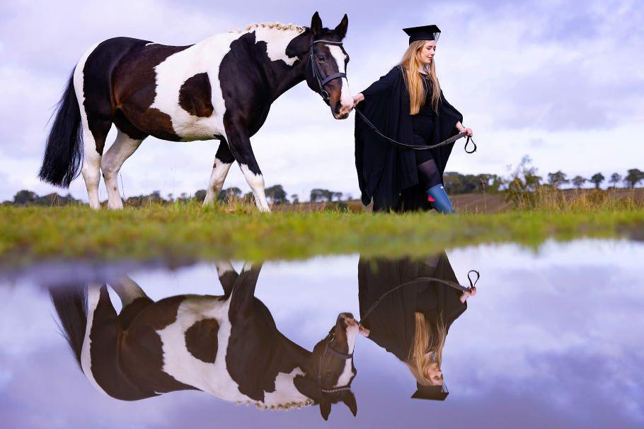 Woman in graduation gown leads a brown and white horse, both reflected in water