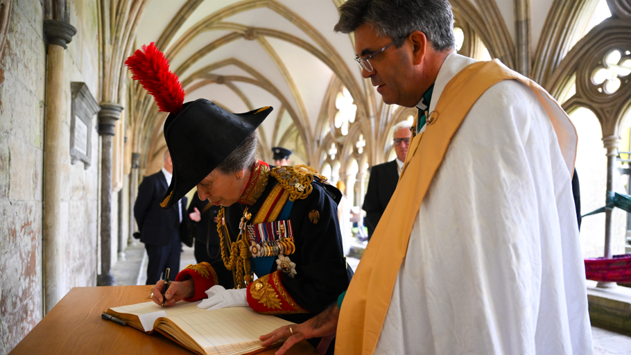 Princess Anne, wearing a military outfit with medals, leans on a wooden table and signs the visitor's book at Salisbury Cathedral as a man in a white cloak watches on.
