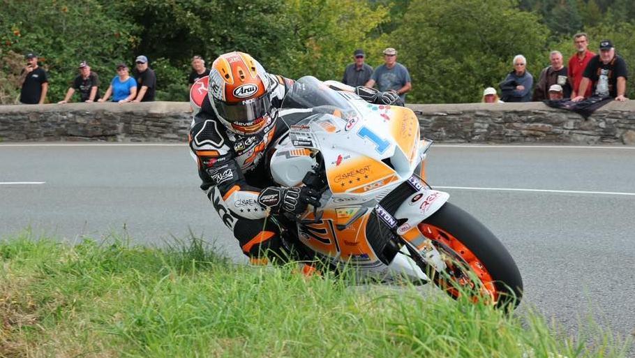 Jamie Williams on his orange white bike that has the number one in blue on the front. His leathers are black with orange and white markings and his helmet is orange and white. He is leaning over on his machine on a bend with spectators watching from behind a low stone wall in the background.