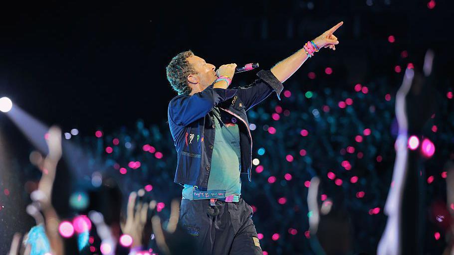 A man standing on a stage with a black microphone in his right hand and his left hand – with colourful wristbands on – pointing in the air. He is wearing black cargo-like trousers, a turquoise blue T-shirt and a black jacket with the sleeves folded up to his elbows. He has short curly brown hair. There are pink and purple lights from the crowd blurred in the background.