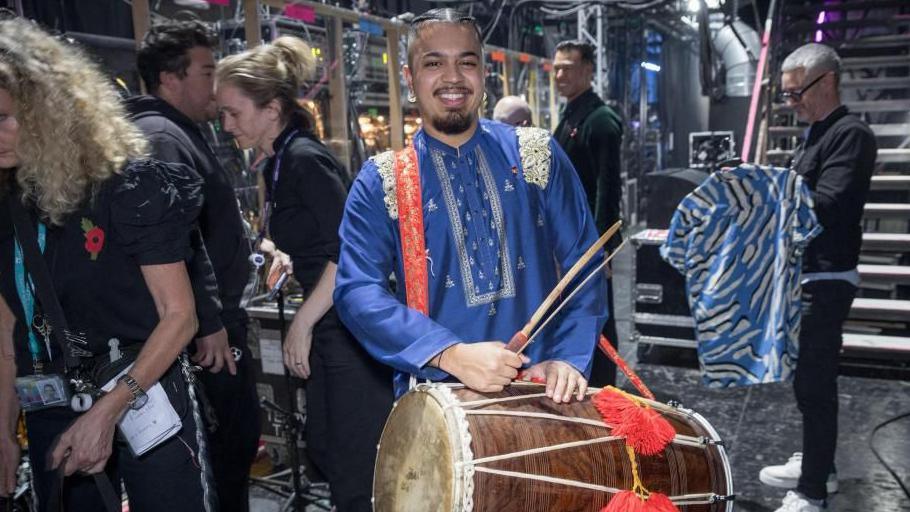 A man standing backstage at a television studio. He is wearing a blue outfit with gold embroidery. He has dark hair that is in two tight plaits. He is holding a large wooden drum with red pom poms. Television crew are in the background, asll wearing black clothes.