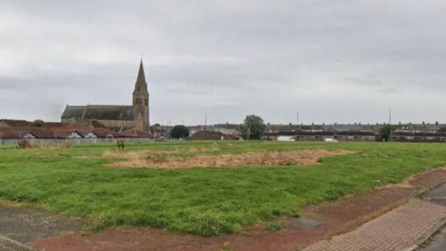 A large field with a brown patch of grass in the middle. There are houses and a church in the background.