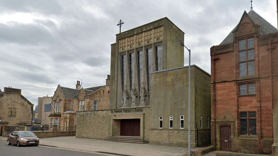 St Francis Xavier Catholic Church in Falkirk. The building is dark stone and has a cross on top of it. A red brick building is to the right and a brown Ford car is parked in front.