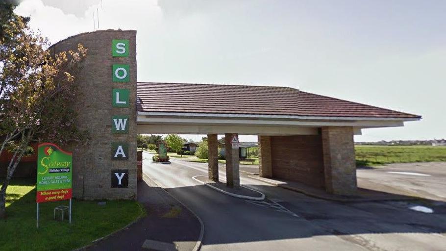 The entrance to Solway Holiday Park. A green sign to the left shows the company's name.  A building with a tiled roof covered the road leading to the building. A stone section of the building contains a vertical green sign which reads Solway.