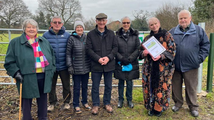 Seven people standing in front of the padlocked entrance to the Glebe. They are all wearing winter coats and hats. Second from the right is Teri Trickett, who is holding the petition that they plan to hand to the Diocese of Lichfield. Behind them you can see a large metal gate, with the Glebe - a green field surrounded by trees and shrubs - beyond the fencing. 