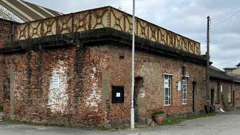 A red brick building showing heavy signs of wear, including white and black patches of paint.  It has black gutters running along the top, and a rusty metal fence on the roof.