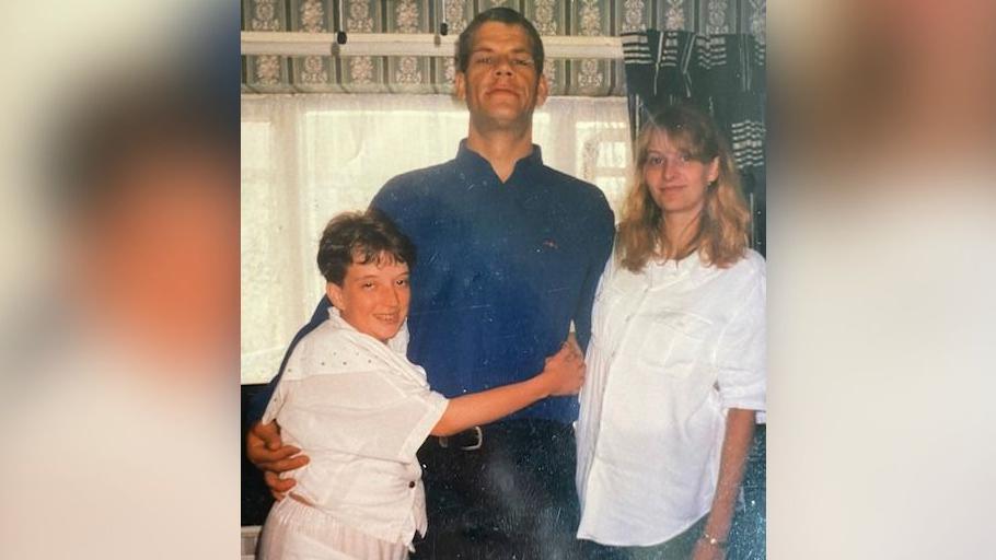 Marina Young pictured smiling and wearing a white blouse and skirt in a house in a family photo. She is hugging her brother Michael, who is smiling and wearing a blue shirt and dark trousers. He is standing next to their sister Michelle, who is also smiling and wearing a white blouse and possibly green trousers, as the photo blurs in this section. 