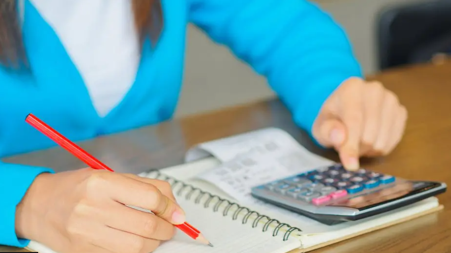 Generic image of a woman in a turquoise cardigan and white t-shirt sitting at a table with a notepad, pencil, calculator and receipts working out her budget and debt.