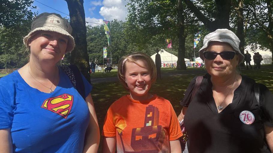 Lynsey is standing on the left of the photo, she has a beige bucket hat on. She is wearing a blue short sleeved top with a classic superman logo in red and yellow. Stood next to her is her daughter Clo who is grinning at the camera. She has short blond hair pushed back with a hair band. She is wearing an orange t-shirt with a Roblox design on. Audrey Lee is stood on the right, she is wearing a white and grey bucket hat, sunglasses and a black t-shirt with a 70s birthday badge pinned to it