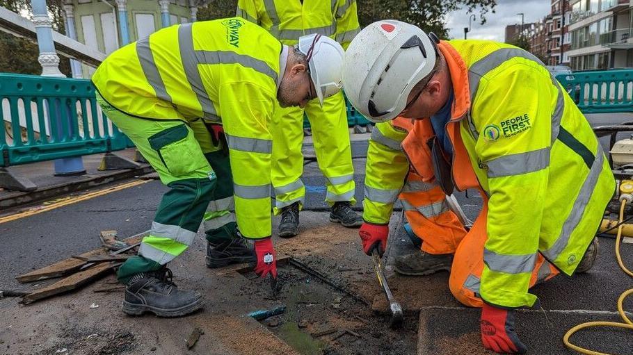 Council workers from People First wearing Hi-Vis and white helmets shown doing some work in the tiles on the ground. In the background, there are turquoise barriers marking the area where the maintenance is taking place.