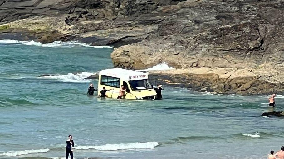 Ice cream van with water above its wheels as people try to stop it going further out to sea