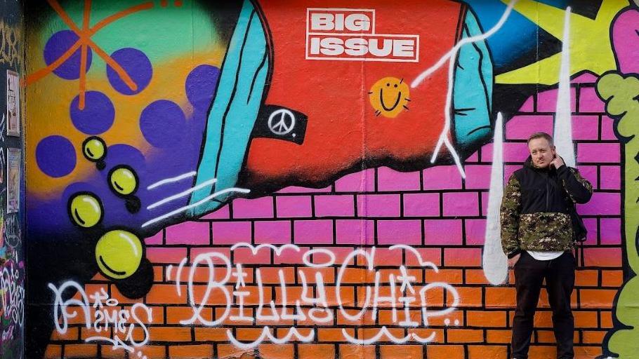 Lewey stands next to his artwork on the mural, featuring bright colours for The Big Issue segment and white spray paint letters that read Billy Chip.