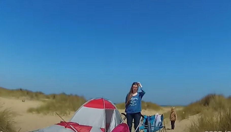 Video grab of a woman and a young child next to a tent on a beach. The woman is wearing a blue top. The child is in the background wearing a brown top and leggings. 