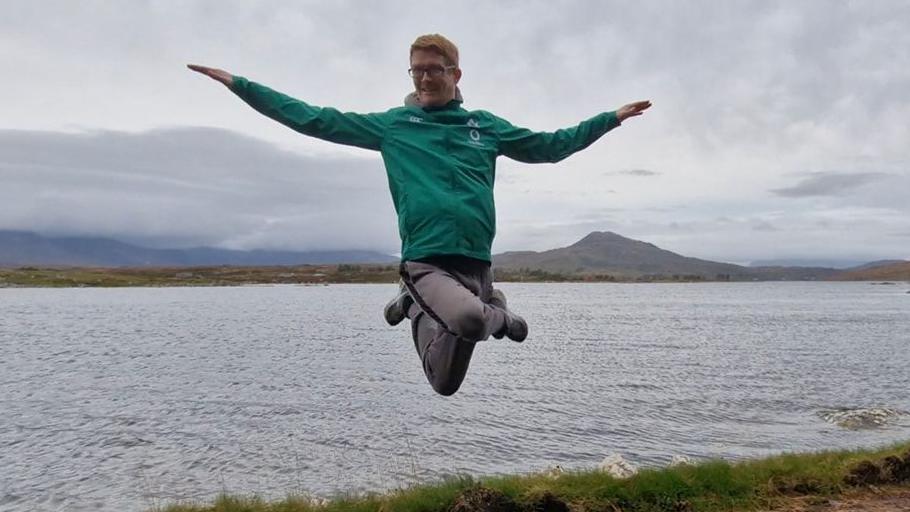 Tom Conroy has been captured in the midst of a dancing move. He appears to be floating in mid air with his legs crossed and arms. Behind him is a vast lake and landscape settings. He has ginger hair, wearing glasses and is wearing a green jacket.