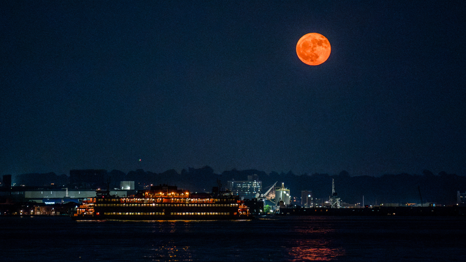 An orange-hued moon hangs in the sky over a river illuminated by lights from nearby properties