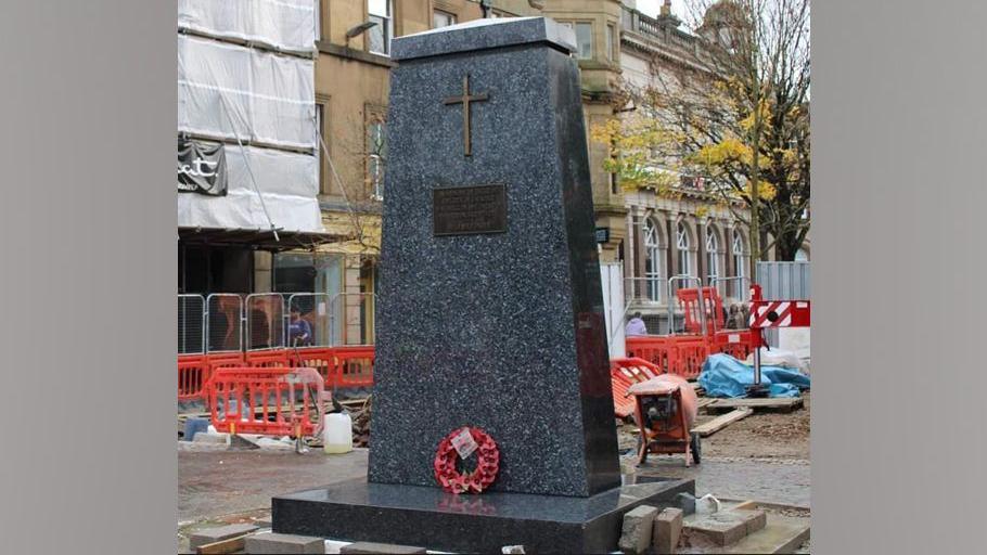 The war memorial in its new location in Market Square. The memorial is a black obelisk, with a cross afixed near its top. There is a red poppy wreath at its base. Building work can be seen taking place behind it.